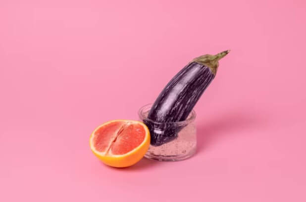 Eggplant standing in a clear glass jar with a sliced orange on a pink background.