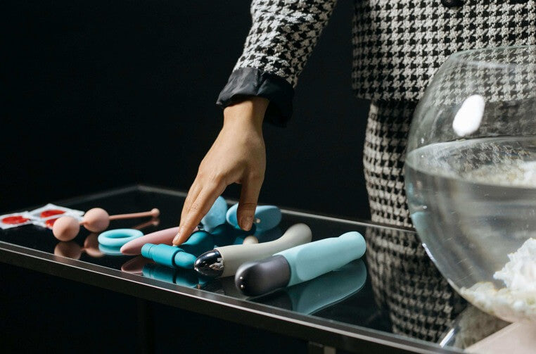 Hand reaching for pastel-colored vibrators and intimate wellness toys on a sleek glass table.