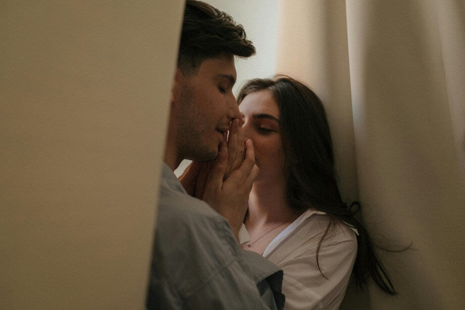Couple sharing a close, romantic moment near soft curtains in warm natural light.