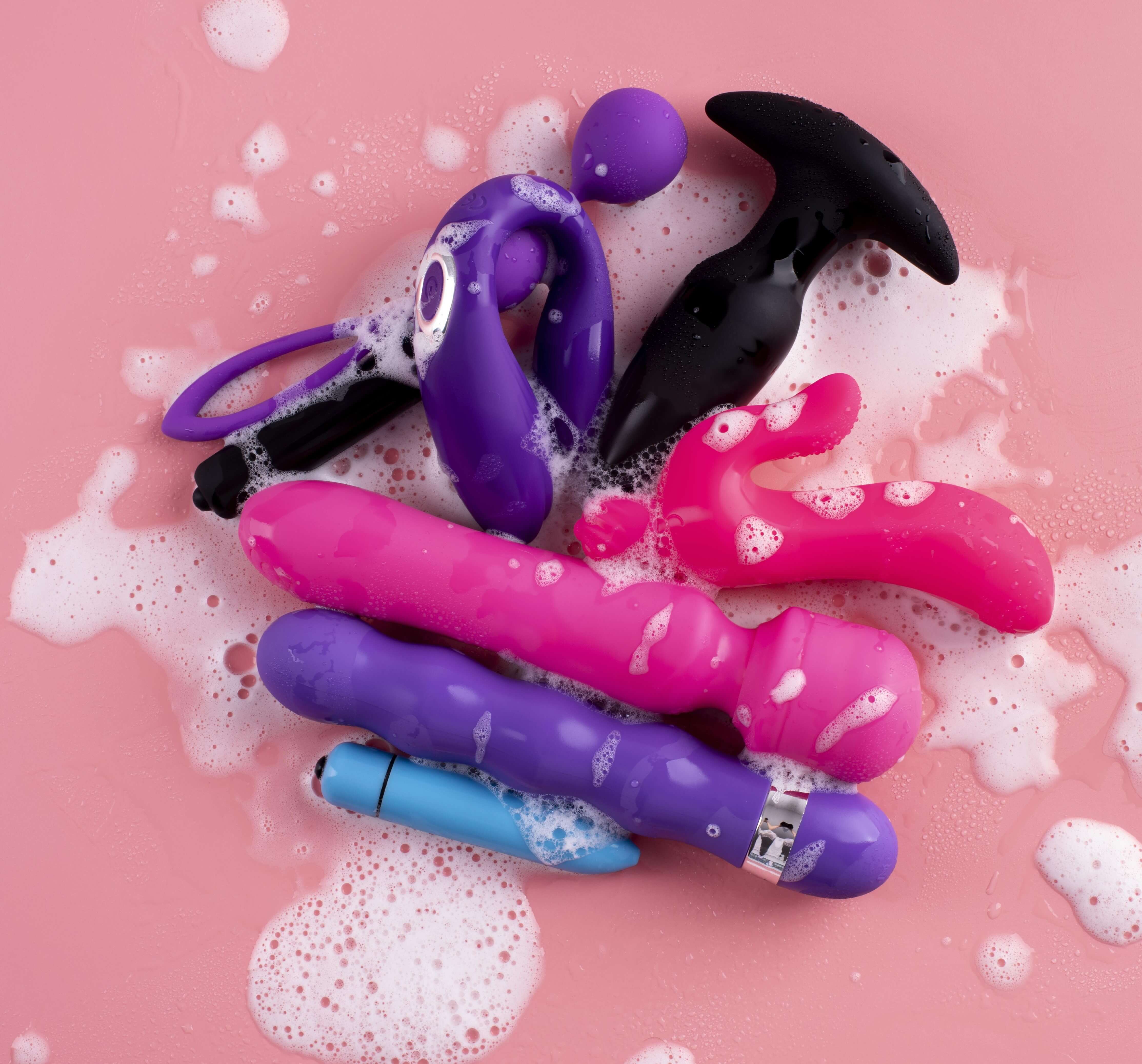 Colorful silicone dildo toys being washed with soap and water on a pink surface, illustrating how to clean silicone dildo products properly for hygiene, comfort, and long-term care.