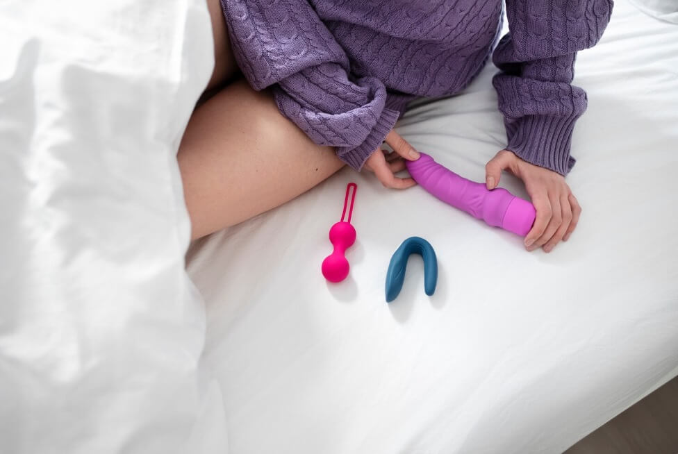 Woman in a purple sweater sitting on bed holding a pink dildo vibrator with other sex toys (Kegel balls and a couple’s vibrator) placed on white sheets – adult toy collection for pleasure and intimacy.