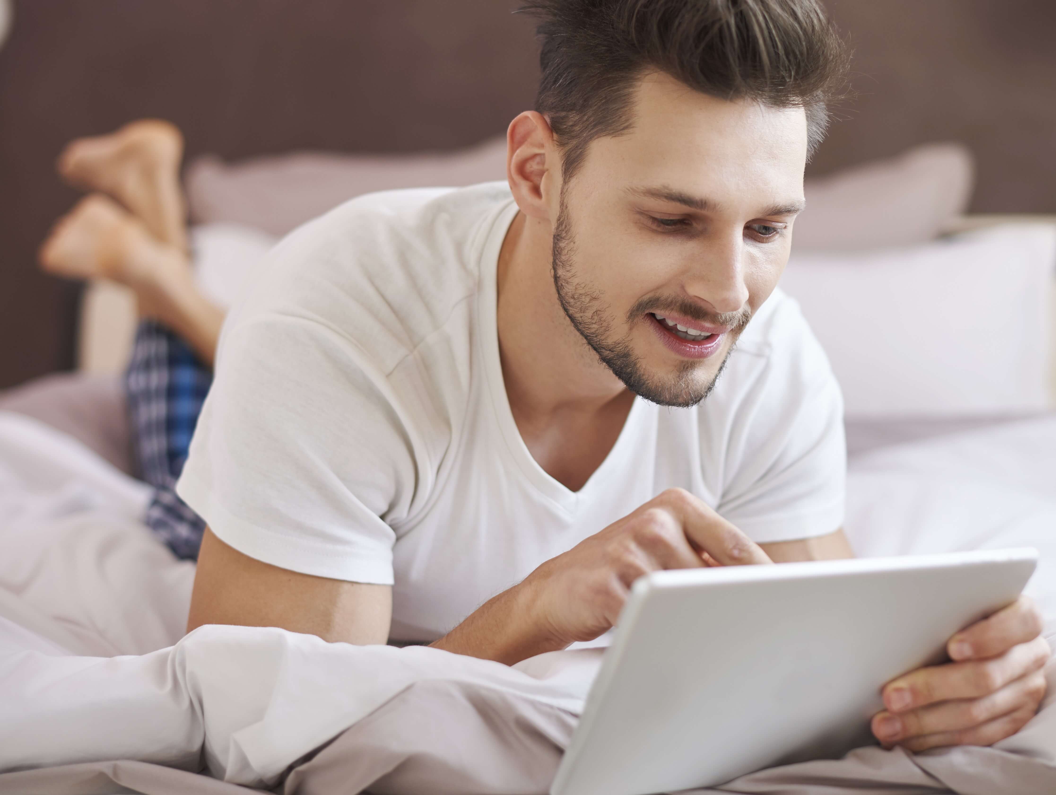 Modern man testing remote-control sex toy technology on his tablet while lying comfortably in bed.
