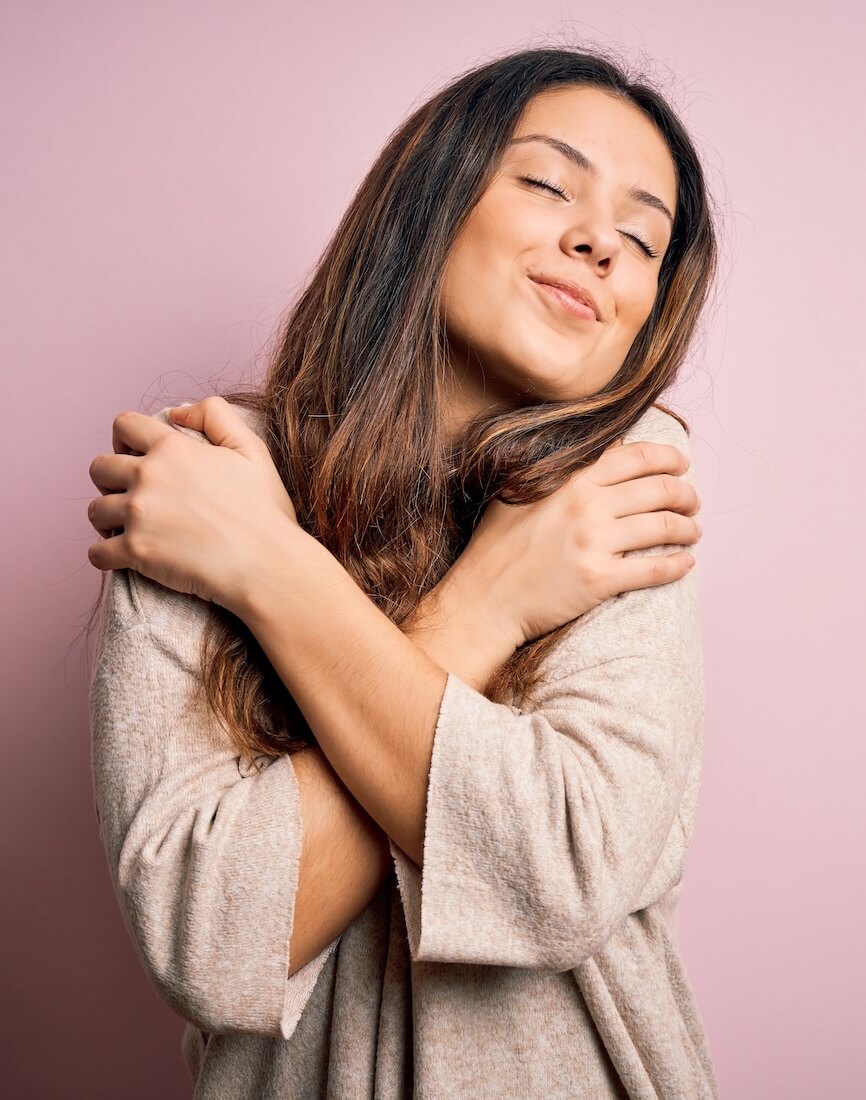 Woman hugging herself for self-love on a pink background after shopping at ThatMood adult toy store.