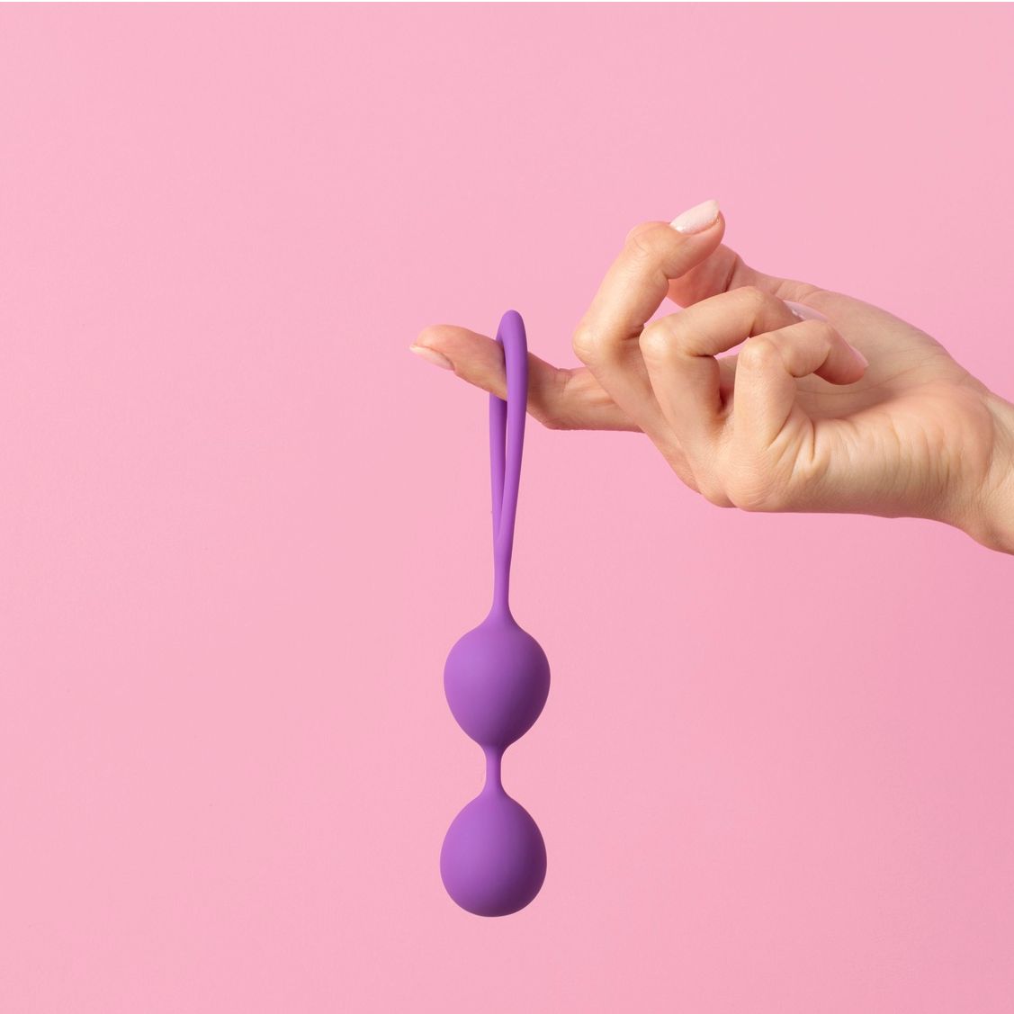 Purple Ben Wa balls hold by a woman finger on a pink background